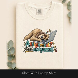 Könnte beinhalten: Ein cremefarbenes T-Shirt mit einer Grafik eines braunen Faultiers, das auf einem Laptop liegt, sowie dem Text "Assignment? What assignment?" in bunten Buchstaben. Das Design enthält kleine "zzz"-Symbole und einen Pfotenabdruck.