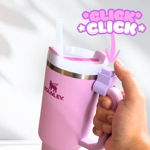 May include: A light pink Stanley tumbler with a white lid, a clear straw, and a matching light pink handle featuring a prominent button mechanism. The text *CLICK* *CLICK* appears in pink with a white outline near the top of the handle.