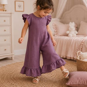Puede incluir: Un mono sin mangas de color morado sólido con mangas con volantes y pantalones de pierna ancha con dobladillos con volantes. La prenda tiene un cuello redondo y una silueta sencilla y holgada que llega hasta la pantorrilla, en tela texturizada.