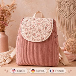 May include: A dusty rose corduroy backpack featuring a rounded flap with a cream-colored floral print. The bag includes a top handle, two side pockets, and adjustable beige straps, showcasing a textured ribbed fabric throughout the main body.