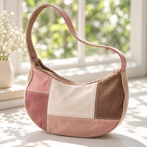 May include: A crescent-shaped shoulder bag constructed from patchwork panels in shades of dusty rose, cream, and dark brown. The bag features a single arched handle, a top zipper closure, and visible white stitching along the seams.