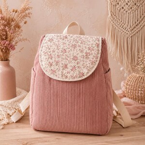 Puede incluir: Una mochila de pana color rosa empolvado con una solapa redondeada con estampado floral sobre fondo crema. La bolsa incluye un asa superior, dos bolsillos laterales y correas de hombro ajustables de tela beige, con textura acanalada.