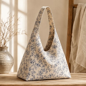 Peut inclure: Un sac hobo triangulaire en tissu blanc cassé orné d'un motif répétitif de vignes florales bleues. Le sac possède une bandoulière intégrée unique qui forme la pointe supérieure de la forme triangulaire, créant une silhouette continue.
