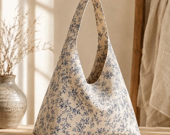 May include: A triangular hobo bag made of off-white fabric featuring a repeating blue floral vine pattern. The bag has a single integrated shoulder strap that forms the top point of the triangular shape, creating a continuous silhouette.