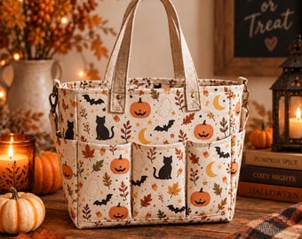 May include: A cream-colored fabric tote bag featuring a repeating Halloween pattern of orange pumpkins, black cats, bats, ghosts, yellow crescent moons, and autumn leaves. The bag has two top handles and three exterior pockets on the front side.
