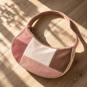 May include: A crescent-shaped handbag featuring a patchwork design of suede-like panels in shades of dusty rose, cream, and dark brown. The bag includes a single shoulder strap and a top zipper closure with visible stitching along the seams.