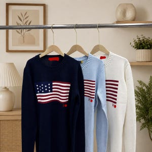 May include: Three long-sleeve crewneck sweaters hanging on wooden hangers. The navy blue sweater features a white and red American flag graphic with stars and stripes. The light blue and white sweaters feature a red and white striped flag graphic.