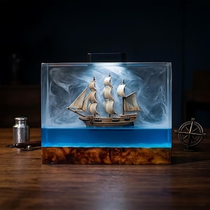 Op de afbeelding: Een rechthoekig harsblok met een houten miniatuur zeilschip met witte zeilen, zwevend in een heldere, wervelende witte mist boven een effen blauwe basis, gemonteerd op een voet van natuurlijk hout met een lamp aan de bovenzijde.