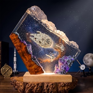 Puede incluir: Una lámpara decorativa de resina que presenta un Halcón Milenario en miniatura y dos cazas estelares X-wing suspendidos dentro de un bloque transparente con temática espacial. La pieza incluye detalles en madera natural, rocas moradas y una base iluminada.