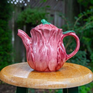 Peut inclure: Une théière en céramique en forme de rose épanouie, avec des pétales rose brillant et un accent de feuille verte sur le couvercle et l'anse. La théière présente un bec verseur incurvé et un corps arrondi, avec une surface texturée imitant les pétales.