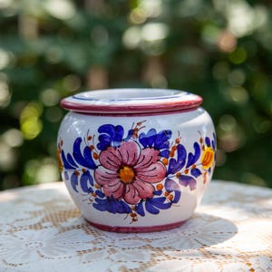 May include: A white ceramic jar with a rounded body and a flat lid. The exterior features hand-painted floral motifs in shades of pink, blue, and yellow, with thin red bands encircling the rim of the lid and the base of the jar.