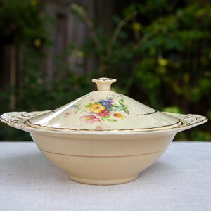 May include: A cream-colored ceramic serving bowl with a matching lid, featuring a floral pattern of yellow, pink, blue, and green flowers on the lid. The bowl has gold-toned trim along the rim and handles, which have a decorative cutout design.