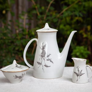 May include: A white porcelain coffee service set consisting of a tall coffee pot with a lid, a lidded sugar bowl, and a small creamer pitcher. Each piece features a delicate grey botanical rose illustration and a thin gold rim trim detail.