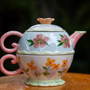 Peut inclure: Un service à thé en céramique pour une personne composé d'une théière bleu clair à motifs floraux roses et d'une tasse assortie ornée de fleurs jaunes. Le couvercle possède une poignée en forme de fleur et les anses sont roses et courbées.