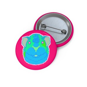 May include: A circular pin-back button featuring a vibrant graphic of a guinea pig face in shades of neon blue, green, and yellow against a bright pink background. The reverse side of the button is metallic silver with a metal pin mechanism.