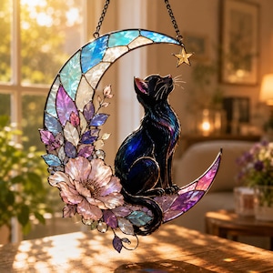 Puede incluir: Un atrapasoles de vidrieras que muestra un gato negro sentado sobre una luna creciente. La luna está compuesta por segmentos de vidrio iridiscente azul, morado y transparente. Una gran flor rosa claro y pequeñas hojas están unidas a la luna, con una pequeña estrella dorada colgando de la punta superior.