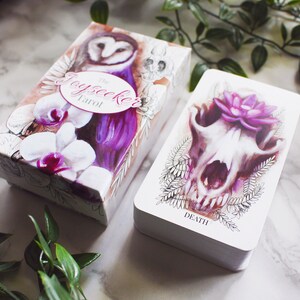 A tarot deck featuring pink and purple animals. Features a barn owl, a corgi puppy, and a wolf skull with a lotus flower. The cards and box are on a white marble background with leaves around them.