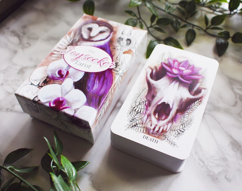 A tarot deck featuring pink and purple animals. Features a barn owl, a corgi puppy, and a wolf skull with a lotus flower. The cards and box are on a white marble background with leaves around them.