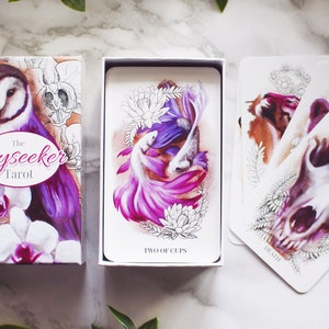 A tarot deck featuring pink and purple animals. Features a barn owl, a corgi puppy, and a wolf skull with a lotus flower. The cards and box are on a white marble background with leaves around them.
