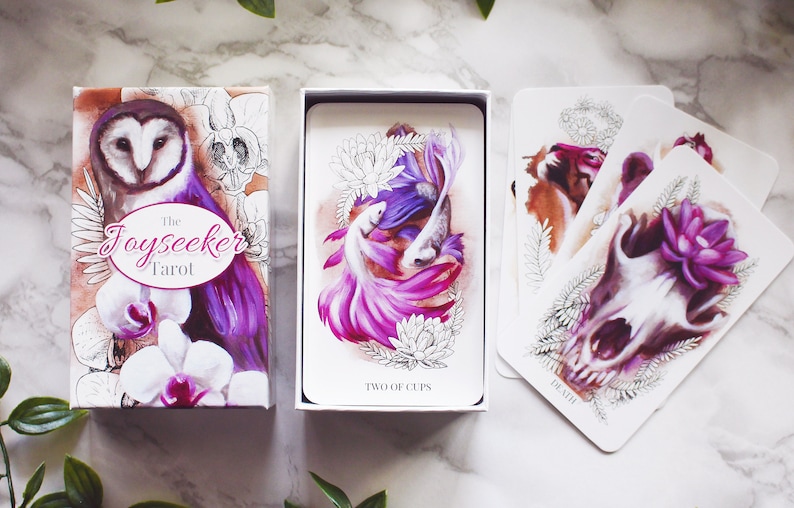 A tarot deck featuring pink and purple animals. Features a barn owl, a corgi puppy, and a wolf skull with a lotus flower. The cards and box are on a white marble background with leaves around them.