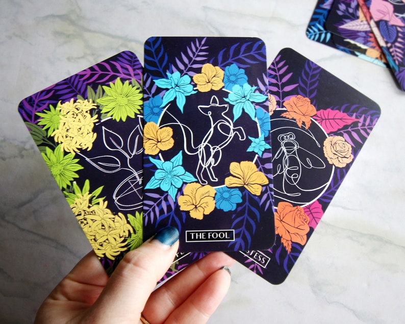 Tarot and oracle cards featuring illustrations on a dark background with brightly colored flowers.