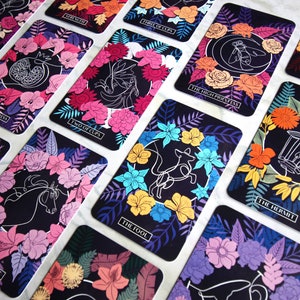 Tarot and oracle cards featuring illustrations on a dark background with brightly colored flowers.