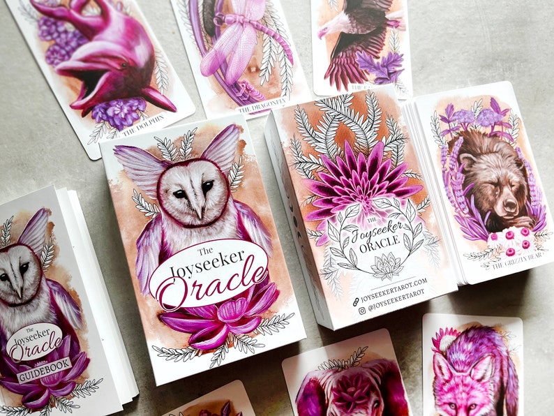 May include: A set of oracle cards with a purple and white color scheme. The cards feature illustrations of animals and nature. The box for the cards has the title "The Joyseeker Oracle" and the website address "joyseekertarot.com".