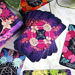 Tarot and oracle cards featuring illustrations on a dark background with brightly colored flowers.