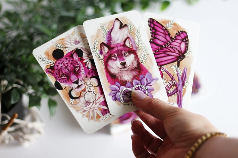May include: Three tarot cards with pink and purple illustrations. The first card shows a pink cheetah with black circles, the second card shows a pink wolf with purple flowers, and the third card shows a pink butterfly with purple flowers.