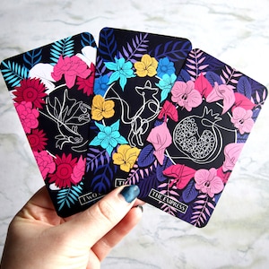 Tarot and oracle cards featuring illustrations on a dark background with brightly colored flowers.