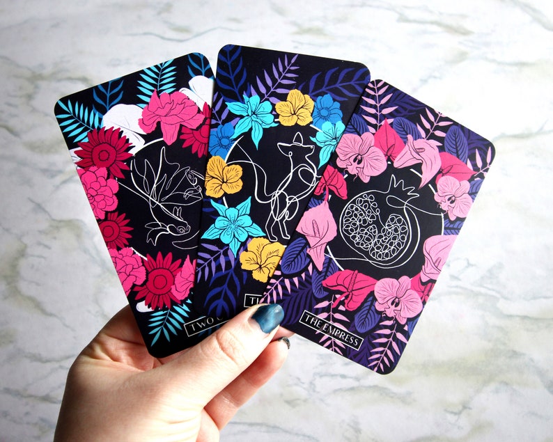 Tarot and oracle cards featuring illustrations on a dark background with brightly colored flowers.