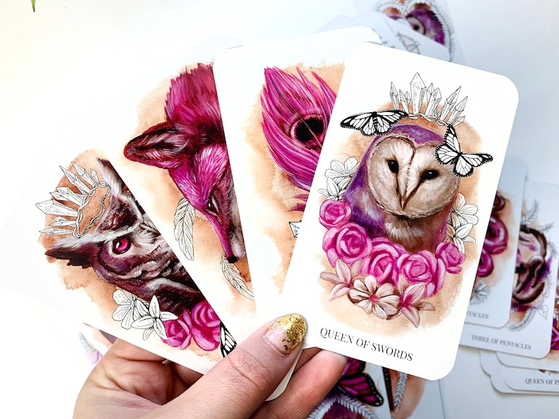A tarot deck featuring pink and purple animals. The cards are being held in a hand, and feature a barn owl with roses