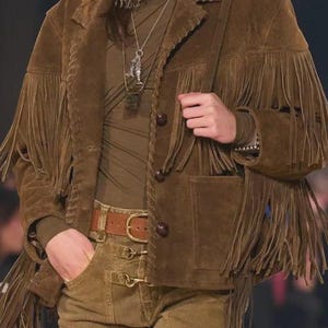 May include: A brown suede jacket featuring long fringe detailing on the sleeves and chest, paired with a brown long-sleeve top, a leopard print scarf, a brown leather belt, and brown trousers with metal hardware accents and a silver fish pendant necklace.