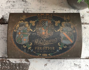 May include: Vintage Whitman's Chocolate box with a gold and black design featuring a crest and the text "Whitman's Prestige Chocolates".