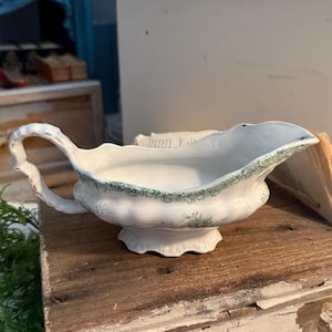 Puede incluir: Salsera de cerámica blanca con borde y asa festoneados, adornada con un delicado estampado floral verde. La salsera se encuentra sobre una superficie de madera envejecida, mostrando su encanto vintage. Una pieza clásica para servir salsas.