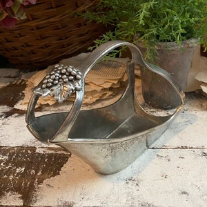 May include: A silver-toned metal wine bottle holder with a handle and grape cluster detail. The basket-shaped holder has a wide opening and a decorative grape vine design. The holder is displayed on a distressed white wooden surface.