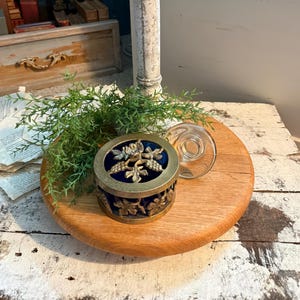 May include: A decorative round wooden tray holds a small, round, antique brass box with a blue velvet interior and a clear glass lid. The box is adorned with a floral design. A faux green plant and a white, distressed pillar are also present.