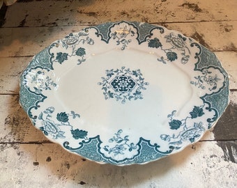 May include: Oval serving platter with a white base and teal floral and scrollwork design. The rim has a scalloped edge. The center features a teal floral medallion. The platter is on a distressed white wooden surface.