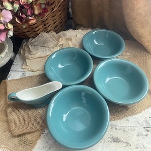 May include: Four teal blue ceramic bowls and a white ceramic gravy boat on a burlap surface. The bowls are all the same size and shape, and they are arranged in a circle. The gravy boat is in the center of the circle.