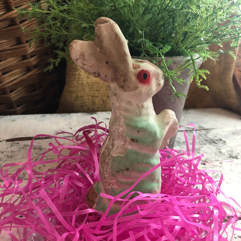 May include: A vintage-style rabbit figurine with a weathered, off-white and pale green finish. The rabbit has prominent red eyes and a textured surface showing signs of age and wear, standing upright in a nest of bright pink shredded paper.