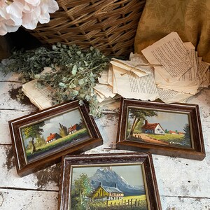 May include: Three framed miniature paintings of countryside scenes. Each painting features a small house, trees, and a mountain in the background. The paintings are in brown frames with a white mat.