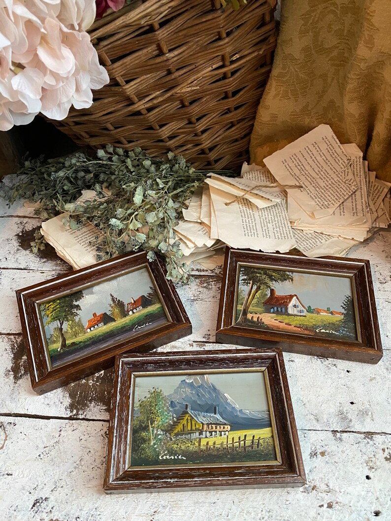 May include: Three framed miniature paintings of countryside scenes. Each painting features a small house, trees, and a mountain in the background. The paintings are in brown frames with a white mat.