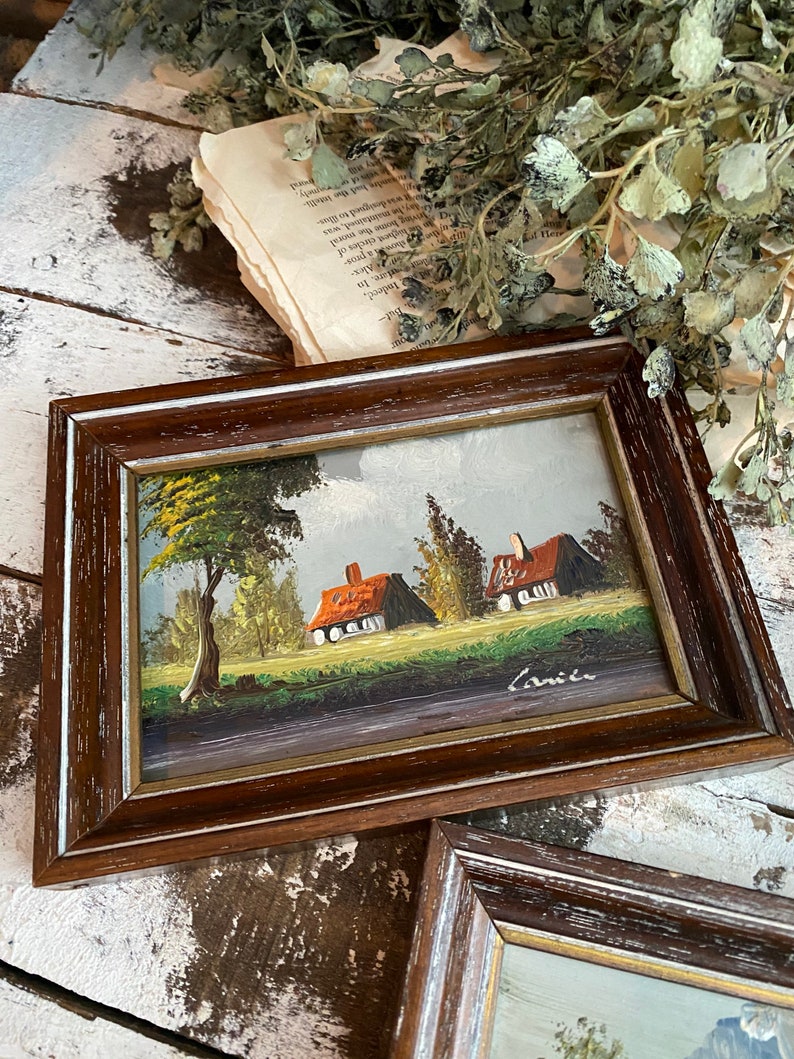 May include: A framed painting of a countryside scene with two houses, a tree, and a blue sky. The painting is signed "Carice" in the bottom right corner.