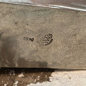 May include: Close-up of a silver metal object with the text "SKS ZINN 95% IN GERMANY" and the number "95%" stamped on the surface. The metal has a textured appearance.