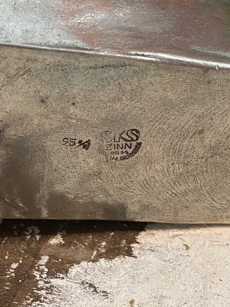 May include: Close-up of a silver metal object with the text "SKS ZINN 95% IN GERMANY" and the number "95%" stamped on the surface. The metal has a textured appearance.