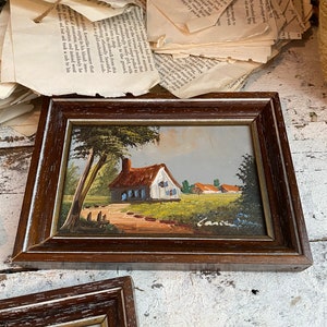 May include: A framed painting of a small cottage with a red roof and blue windows, surrounded by trees and a path leading to the front door. The painting is set against a backdrop of a blue sky and green grass.