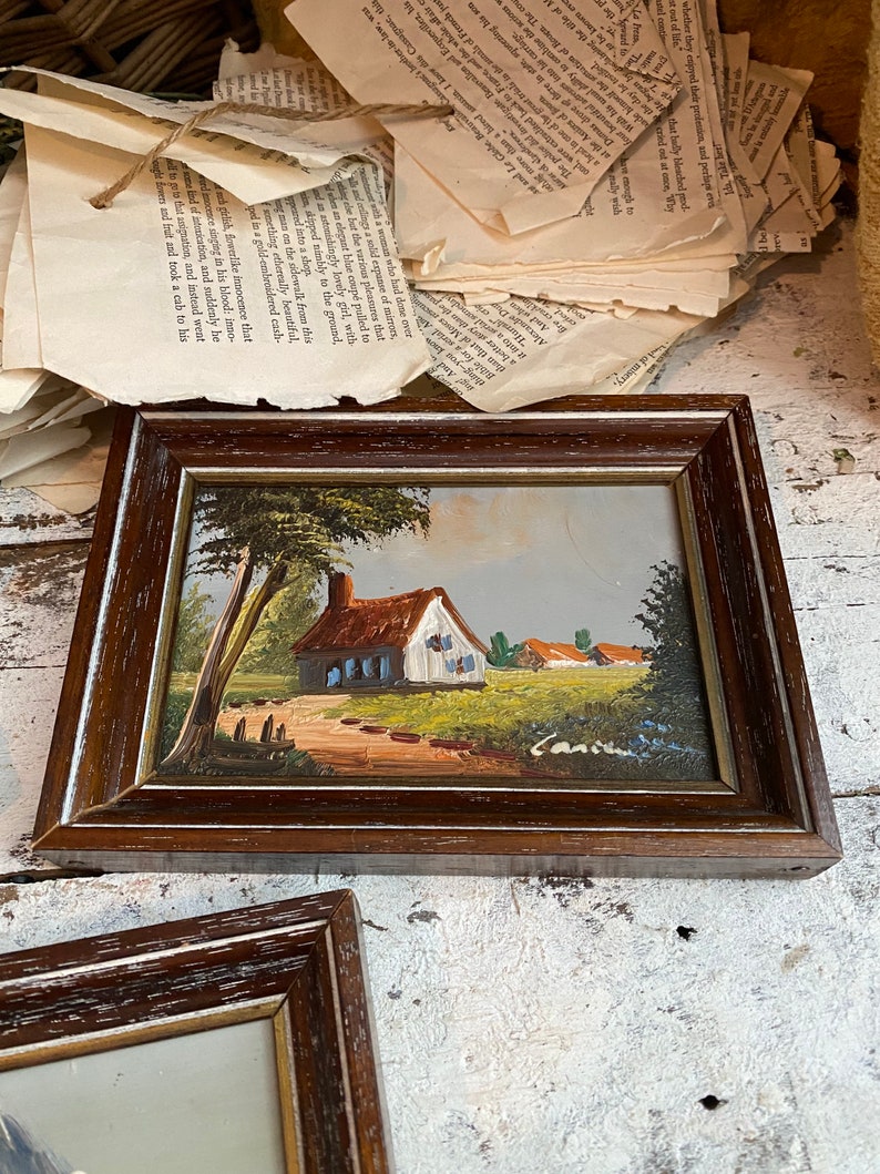 May include: A framed painting of a small cottage with a red roof and blue windows, surrounded by trees and a path leading to the front door. The painting is set against a backdrop of a blue sky and green grass.