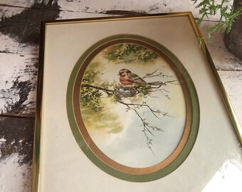 May include: A framed watercolor painting of a bird sitting on a nest in a tree. The painting is surrounded by a double oval frame with a gold and green border.