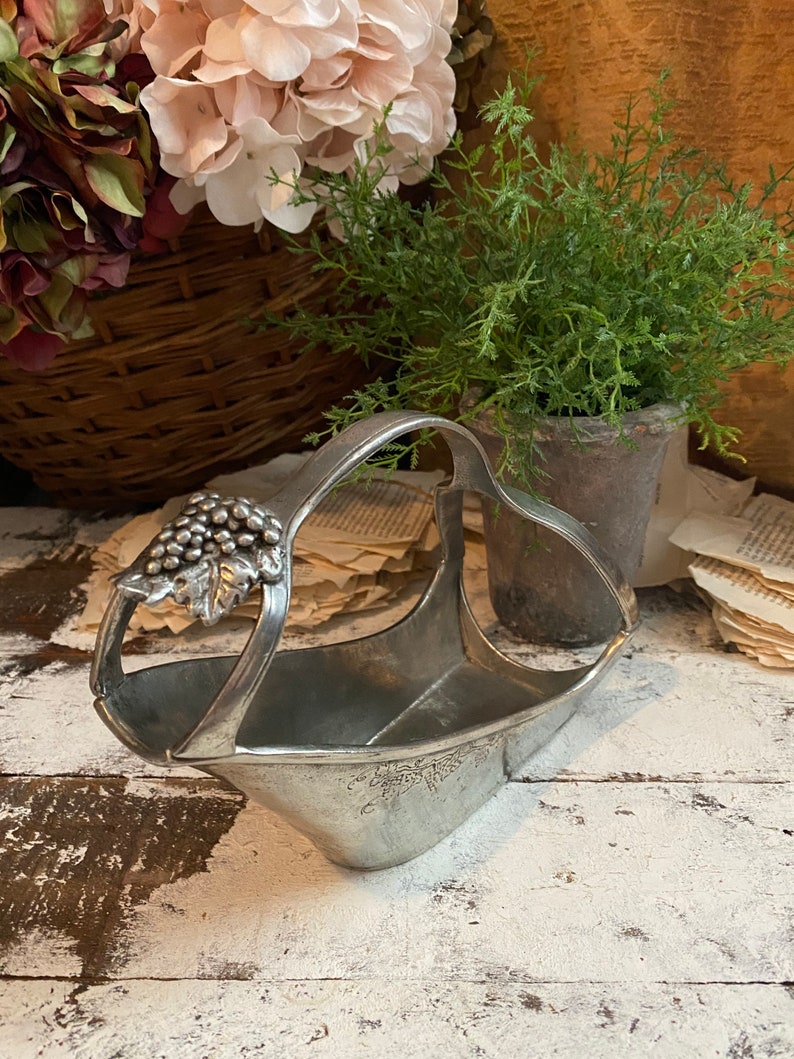 May include: A silver-toned metal wine bottle holder with a handle and grape cluster detail. The basket-shaped holder has a wide opening and a decorative grape vine design. The holder is displayed on a distressed white wooden surface.