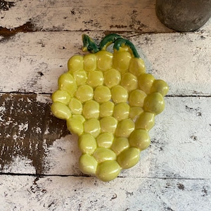 May include: A ceramic grape dish with a green stem and leaves. The dish is shaped like a bunch of grapes and is a light yellow color.